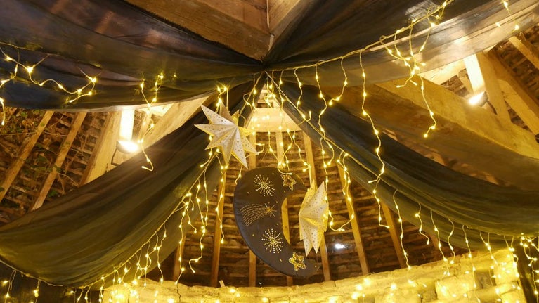 The timber frame ceiling of Avebury's 16th Century Dovecote decorated with black drapes, hanging paper star ornaments, warm fairy lights, and a crescent moon decoration featuring golden star and comet designs.